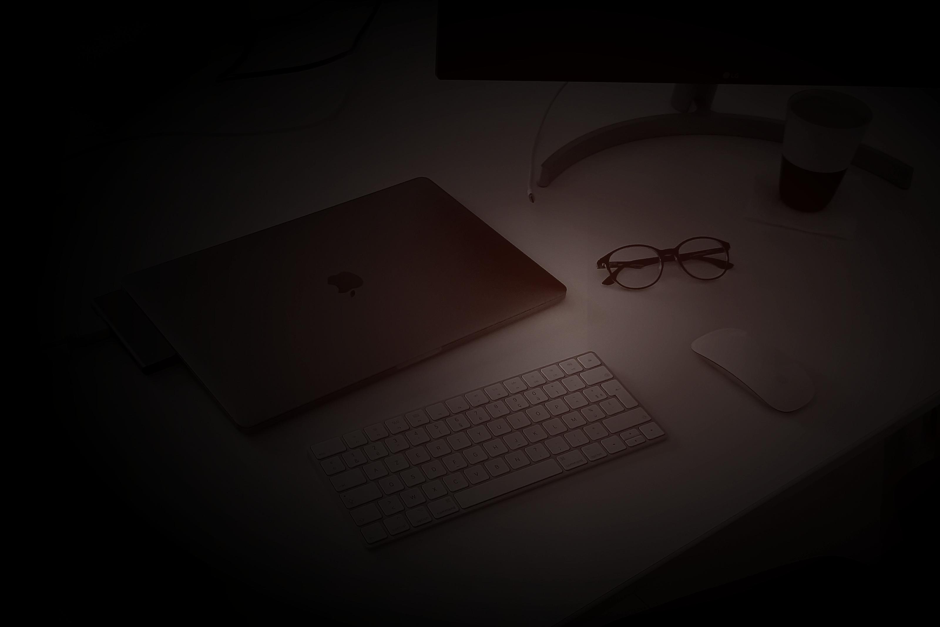 A laptop, keyboard, glasses, and coffee cup on a desk lit by a single lamp at night